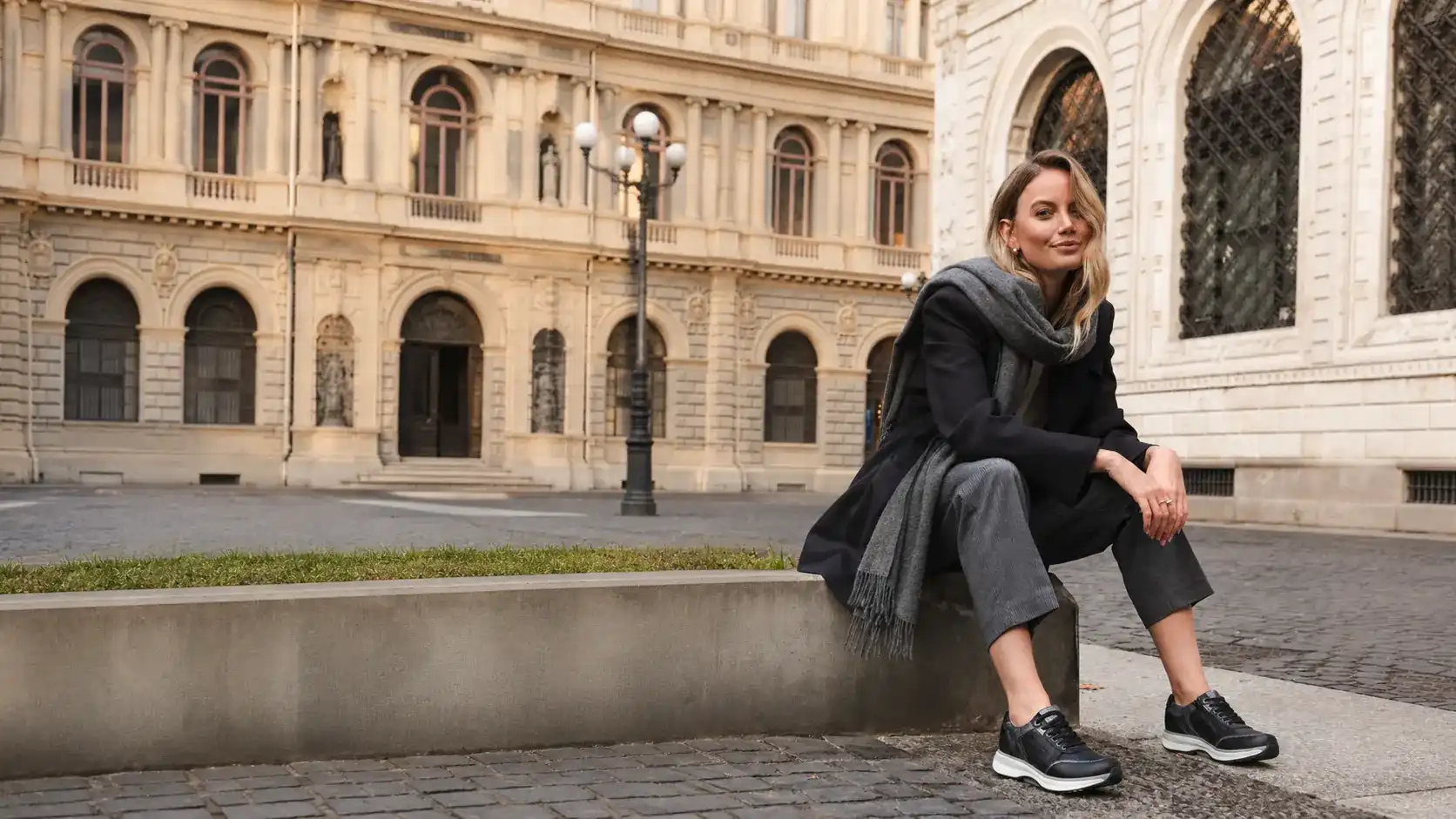 Frau sitzt auf einer Mauer vor einem historischen Stadtgebäude und trägt schwarze Sneaker mit heller Sohle