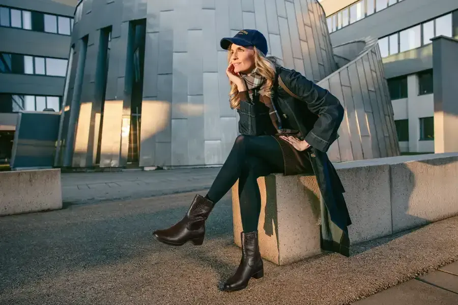 Frau mit Basecap und Stiefeletten sitzt in der Abendsonne vor moderner Architektur
