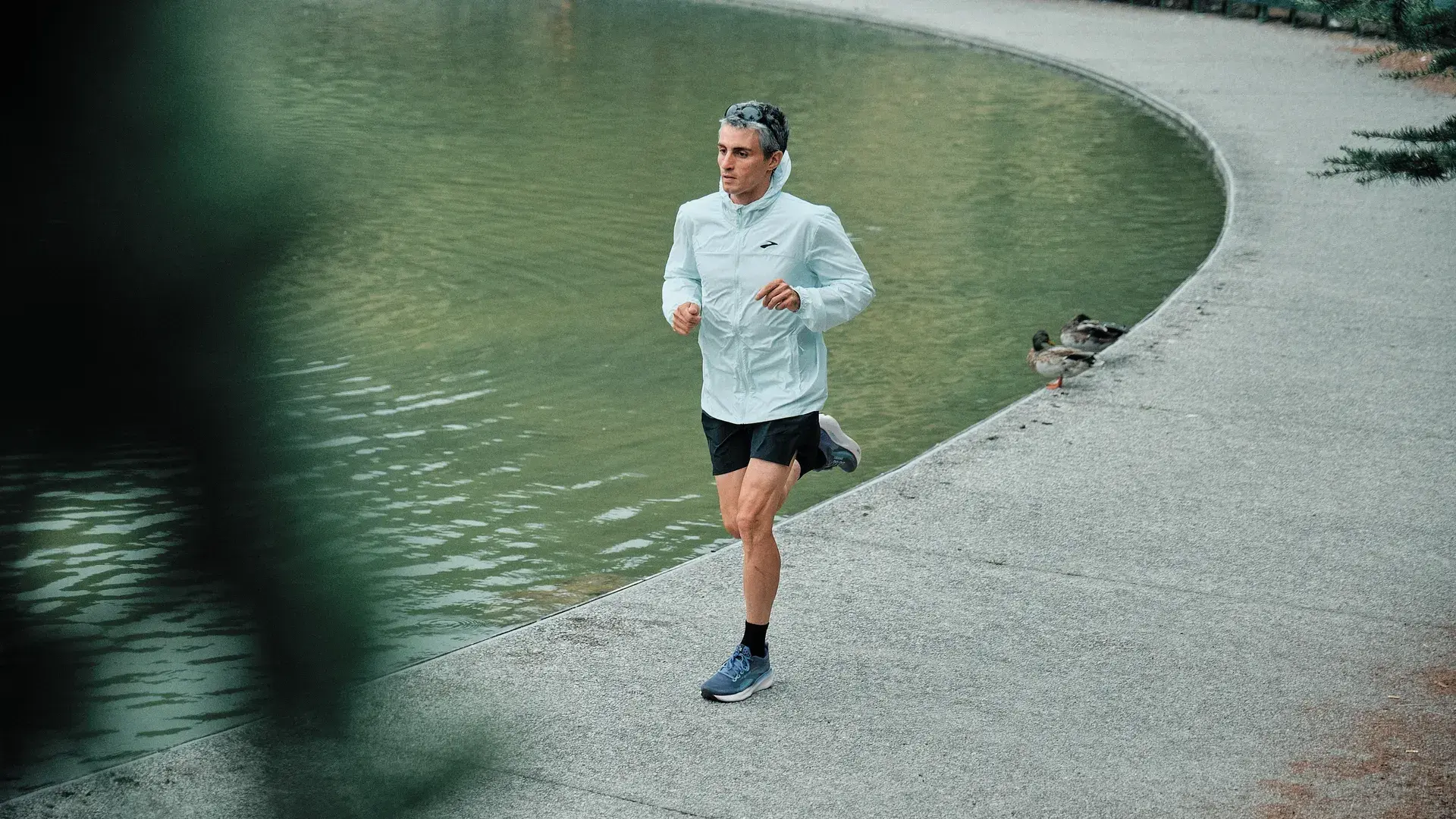 Läufer in heller Laufjacke und blauen Brooks-Laufschuhen auf einem Parkweg am Teich